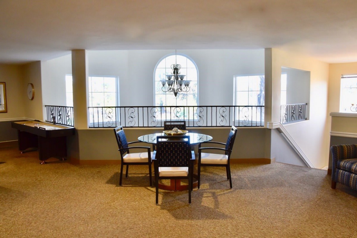 A spacious dining area with a round table and a chandelier, featuring large windows and a carpeted floor.