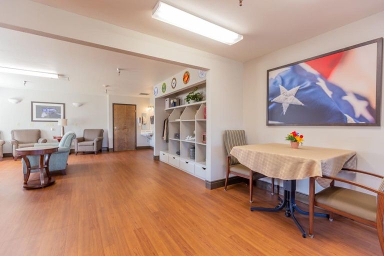 A well-lit room with a table, chairs, and shelves, featuring artwork on the wall.