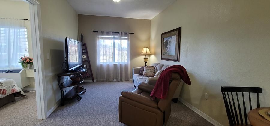 A cozy living room with a couch, recliner, and TV, warmly lit by a lamp near the window.