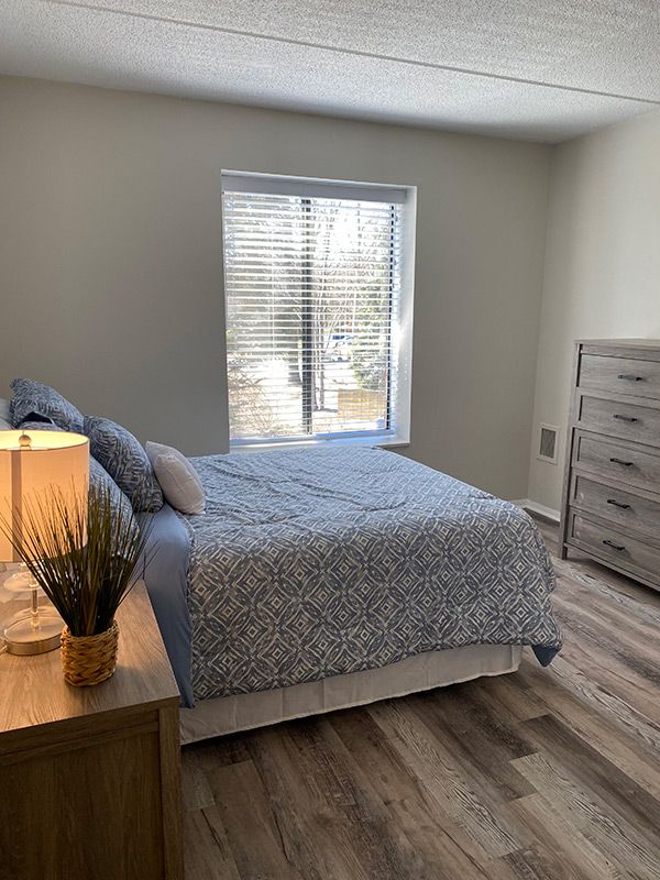 A cozy bedroom with a neatly made bed, wooden dresser, and a lamp on the nightstand.