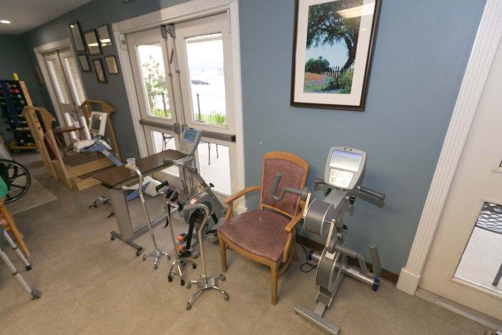 A room with exercise equipment including stationary bikes and a chair, featuring a painting on the blue wall and windows in the background.