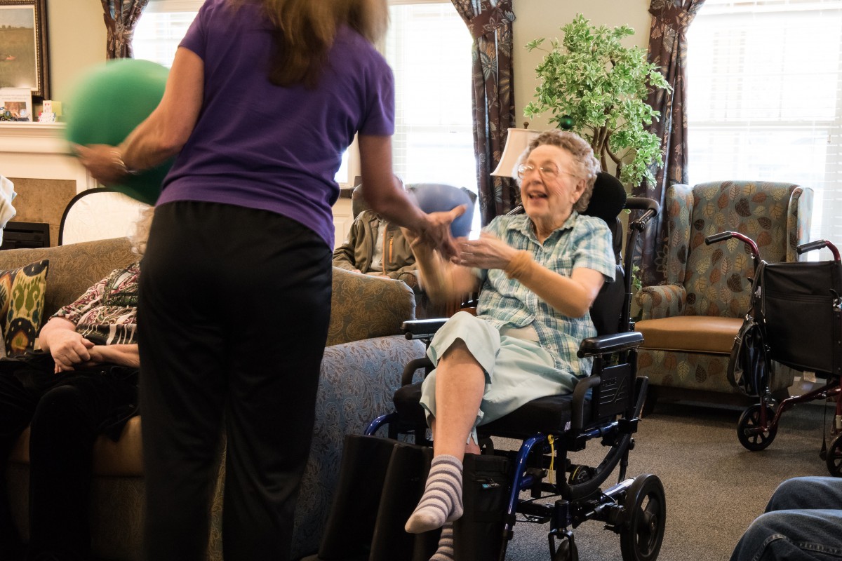An elderly person in a wheelchair shares a joyful moment with a caregiver indoors.