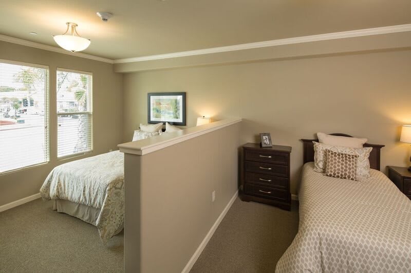 A cozy bedroom featuring two beds separated by a partial wall, decorated with neutral tones and well-lit by natural and artificial light.
