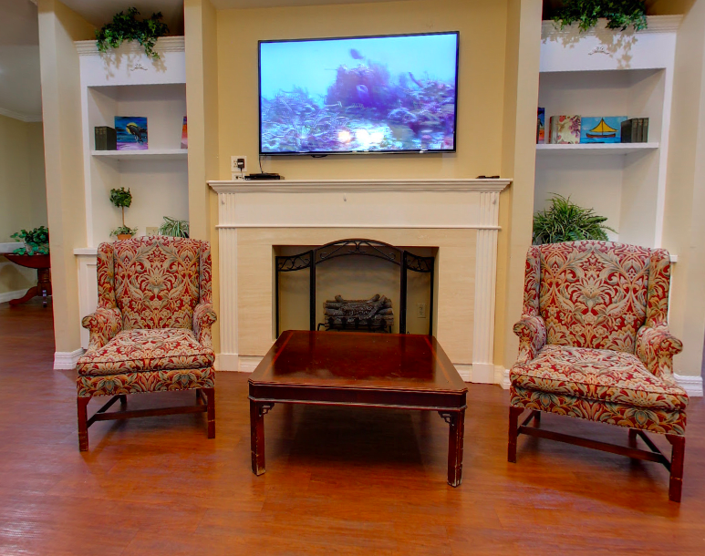 A cozy living room with a television above the fireplace, flanked by bookshelves and armchairs.