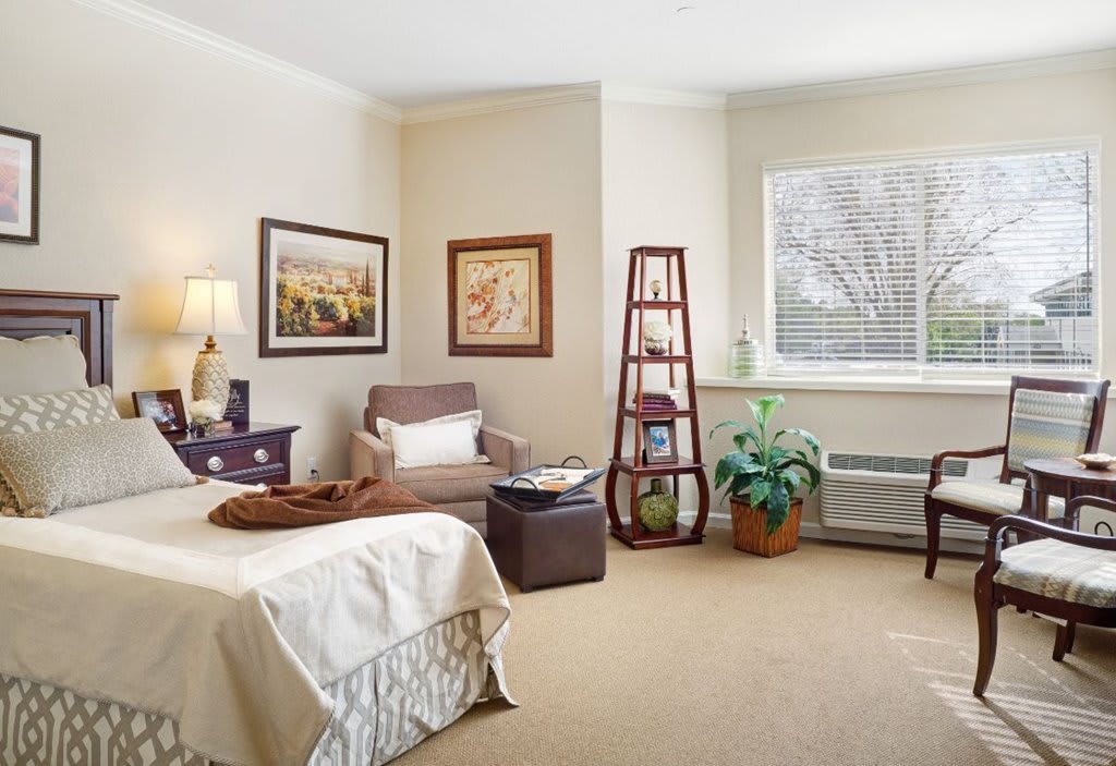 A cozy bedroom interior with a bed, armchair, artwork, and decorative plants.
