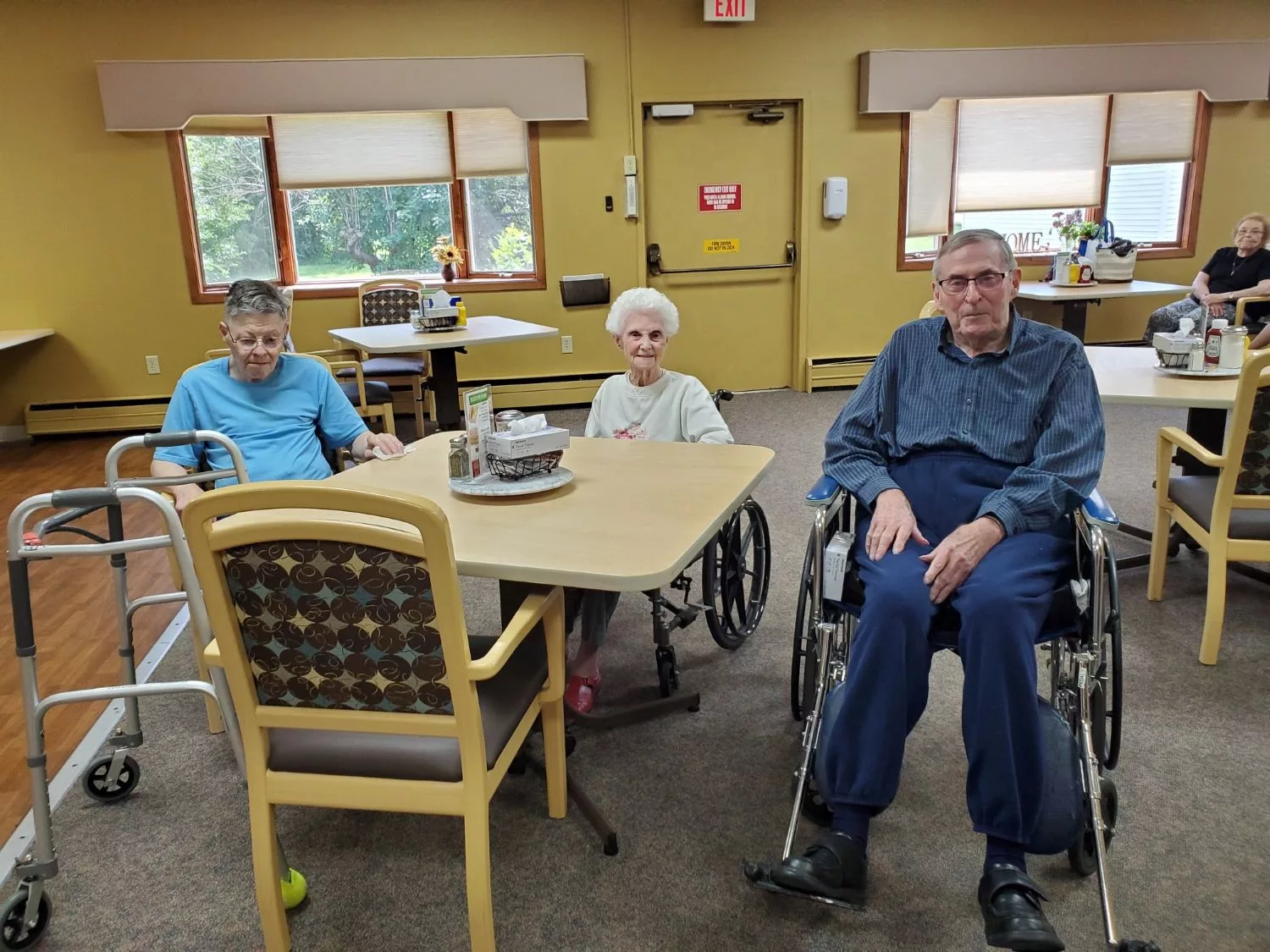 Three elderly individuals sitting together in wheelchairs at a table in a community room.