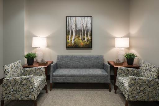 A cozy waiting area featuring a grey sofa, patterned chairs, decorative lamps, and artwork of trees.
