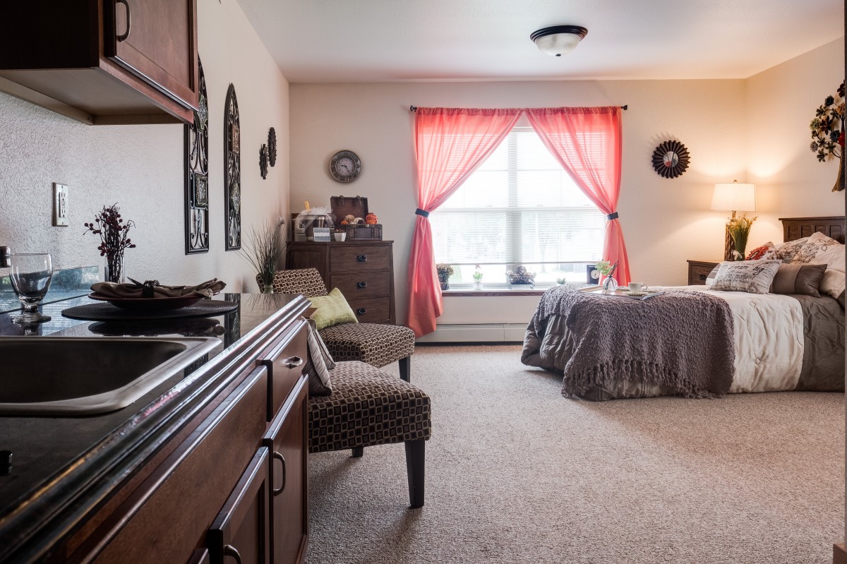A cozy bedroom interior with a bed, dresser, and seating area, featuring natural light through bright curtains.