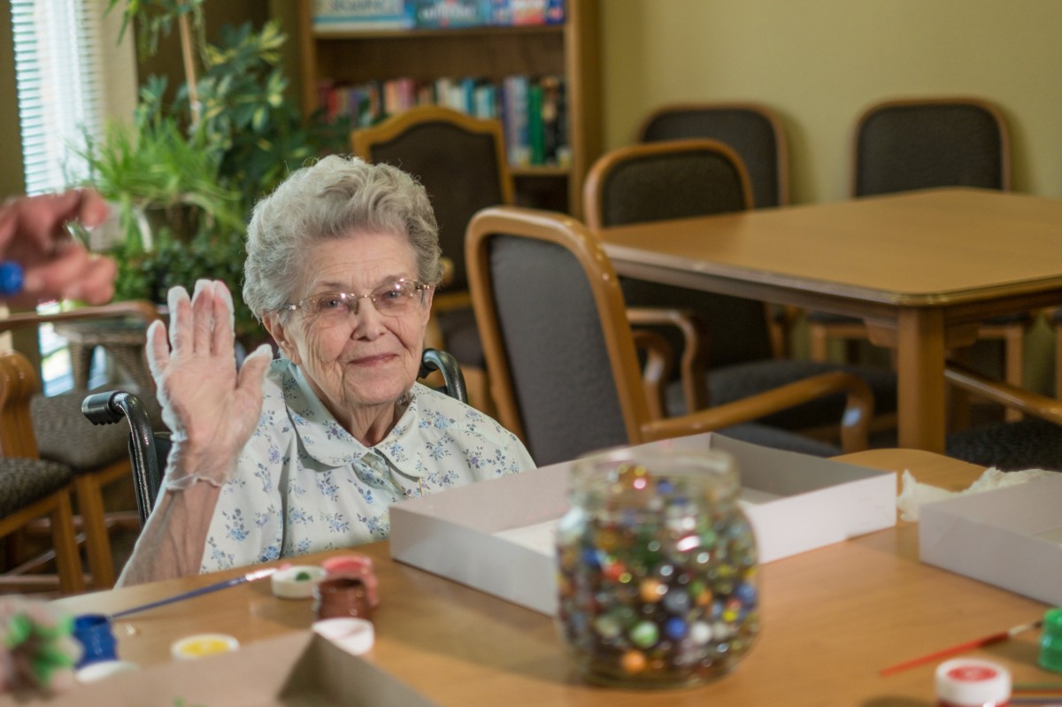 An elderly woman waves while seated at a table in a senior living room.