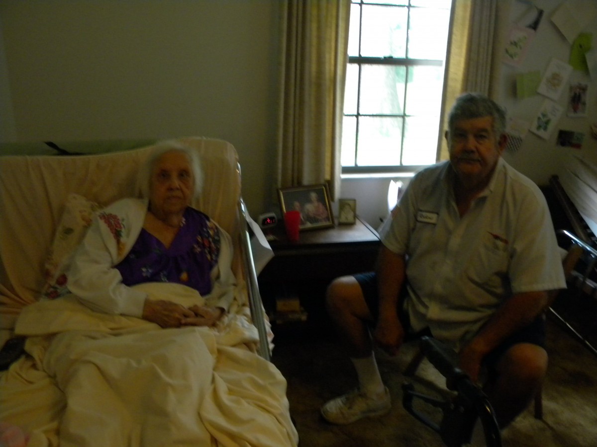 An elderly woman sitting in bed with a man seated beside her in a cozy room.