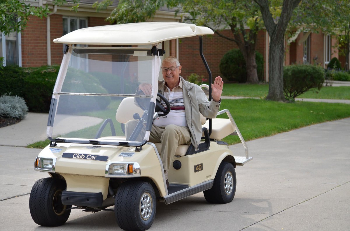 An elderly person is driving a golf cart and waving while on a driveway.