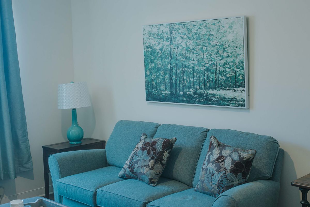 A cozy living room with a teal sofa, artistic cushions, and a matching lamp next to a forest-themed painting.