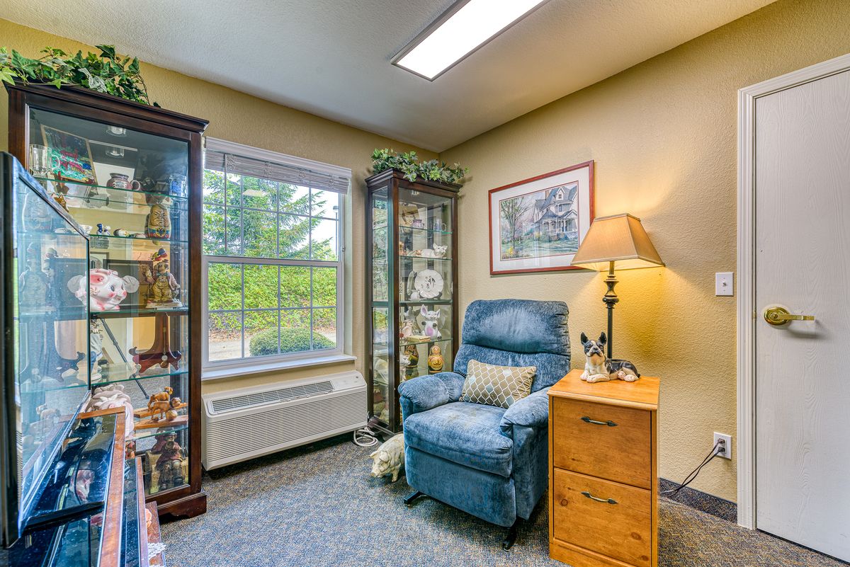 A cozy corner of a living room featuring a blue recliner chair beside a wooden side table with a lamp, surrounded by two glass cabinets displaying decorative items and an indoor plant near the window.