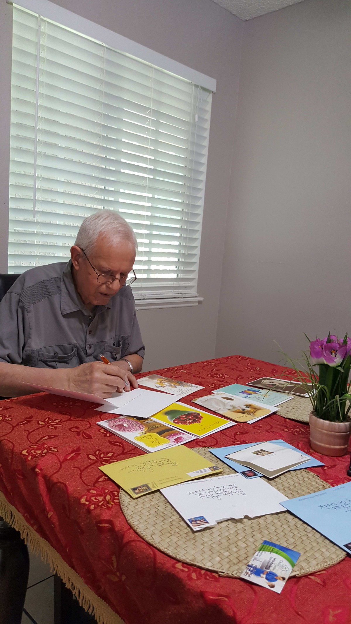 An elderly man writing cards at a table with flowers and several greeting cards.
