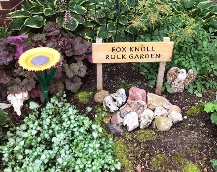 A small rock garden with a sign reading 'Fox Knoll Rock Garden', surrounded by various plants.