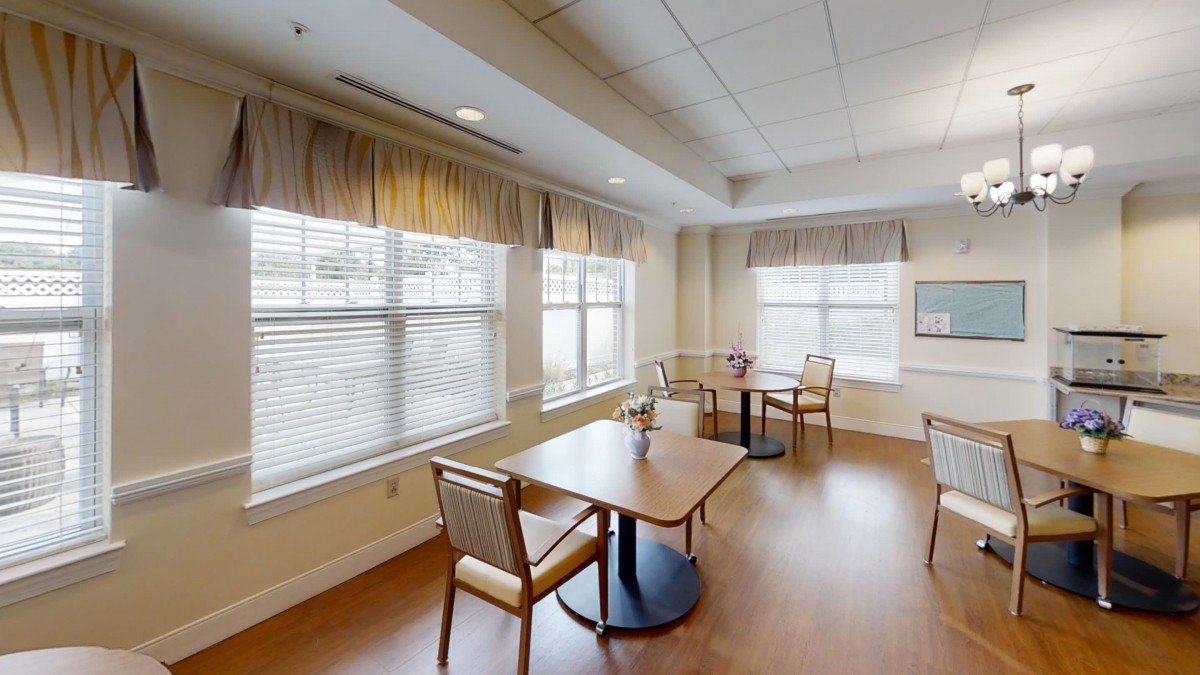 A bright dining room with tables and chairs, featuring large windows and curtains.
