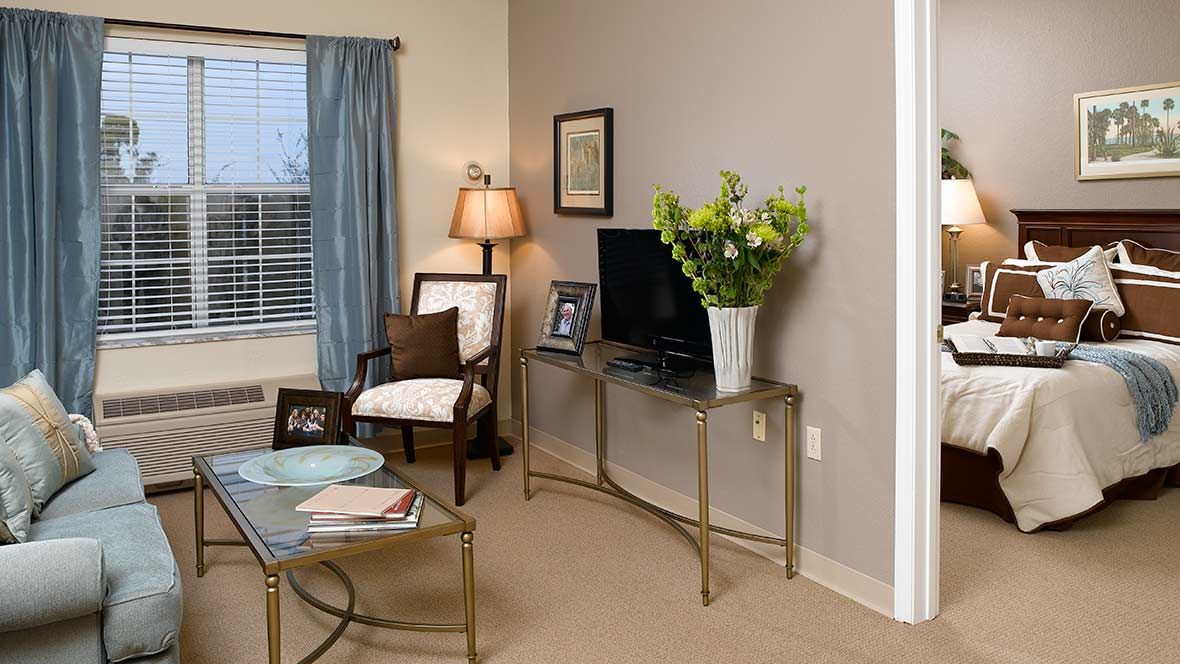 A cozy living room with elegant decor, featuring a TV on a glass table, a lamp, flowers, and comfortable seating.