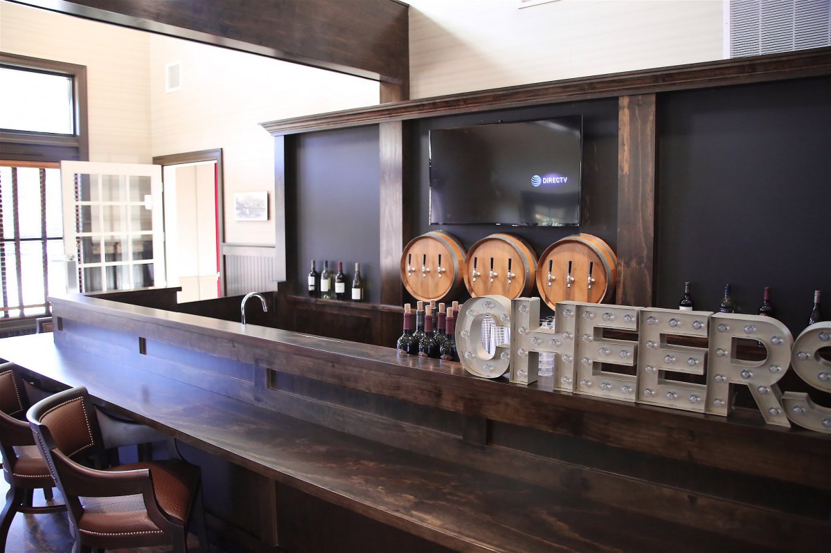 A rustic bar interior featuring a wooden counter, wine bottles, and decorative barrels.
