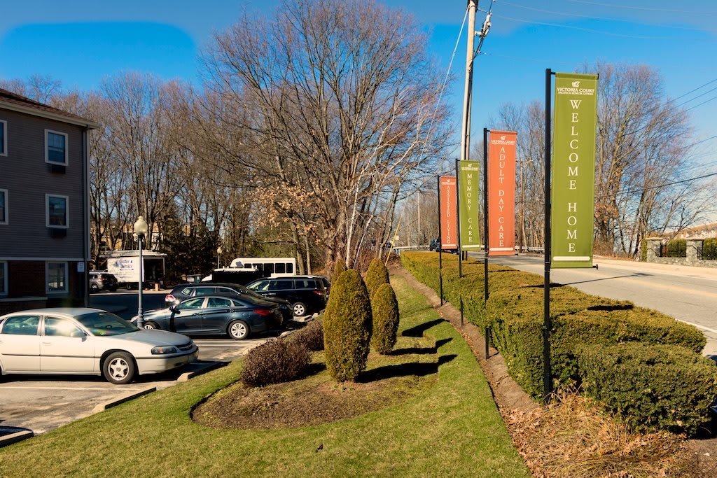 A parking lot beside a road with colorful banners and neatly trimmed shrubs on a sunny day.