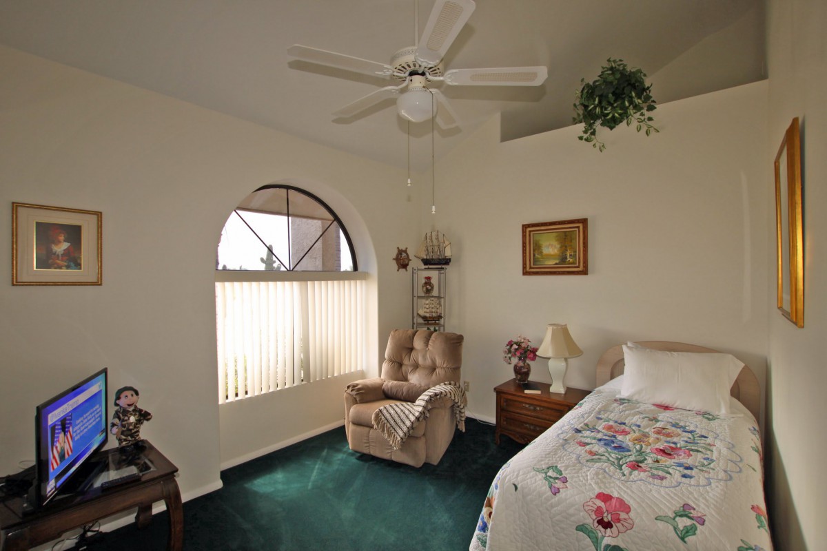 A cozy bedroom with a green carpet, featuring an armchair by a window, a ceiling fan, and floral bedding.