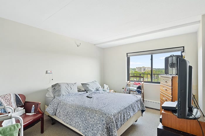A bright bedroom with a bed, dresser, and window overlooking greenery.