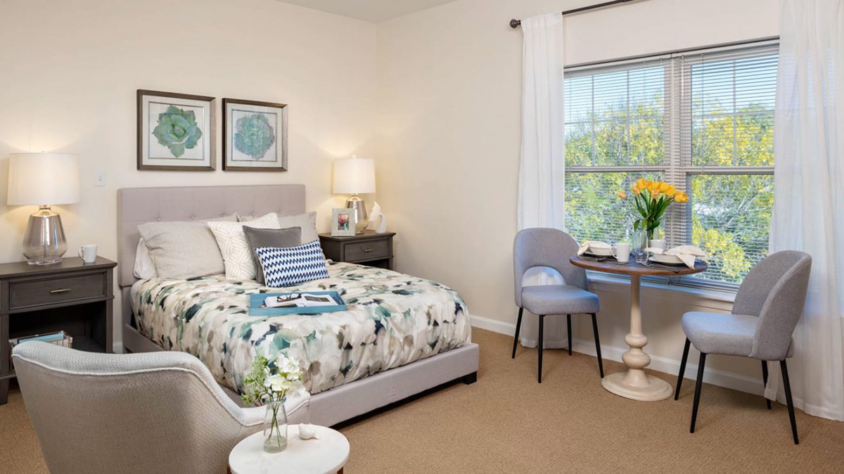 A cozy bedroom with a neatly made bed, bedside lamp, small round table with a chair by the window, and decorative elements creating a tranquil atmosphere.