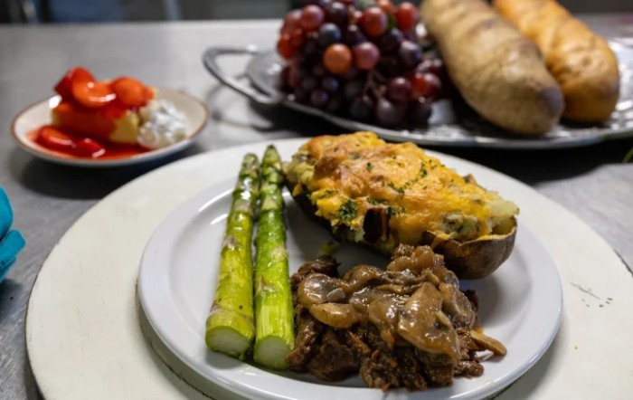 A plated meal featuring grilled asparagus, stuffed potato, and sauted mushrooms, with grapes and bread in the background.