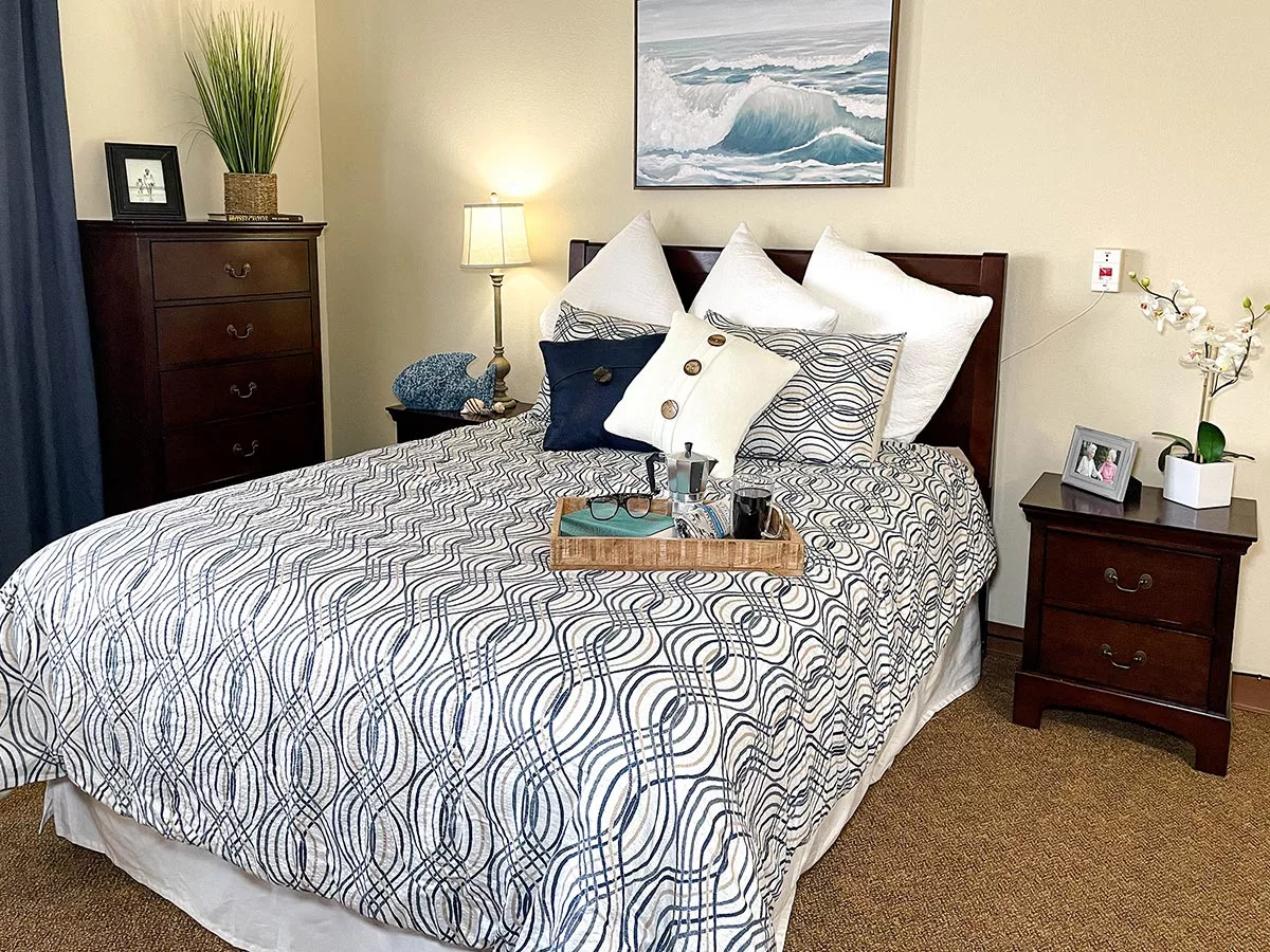 A neatly arranged bedroom featuring a patterned bedspread, decorative pillows, and a bedside table with a lamp and artwork on the wall.