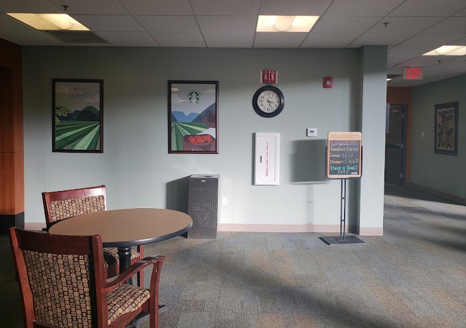 A quiet waiting room with chairs and a table, adorned with paintings on the walls and a clock above a notice board.