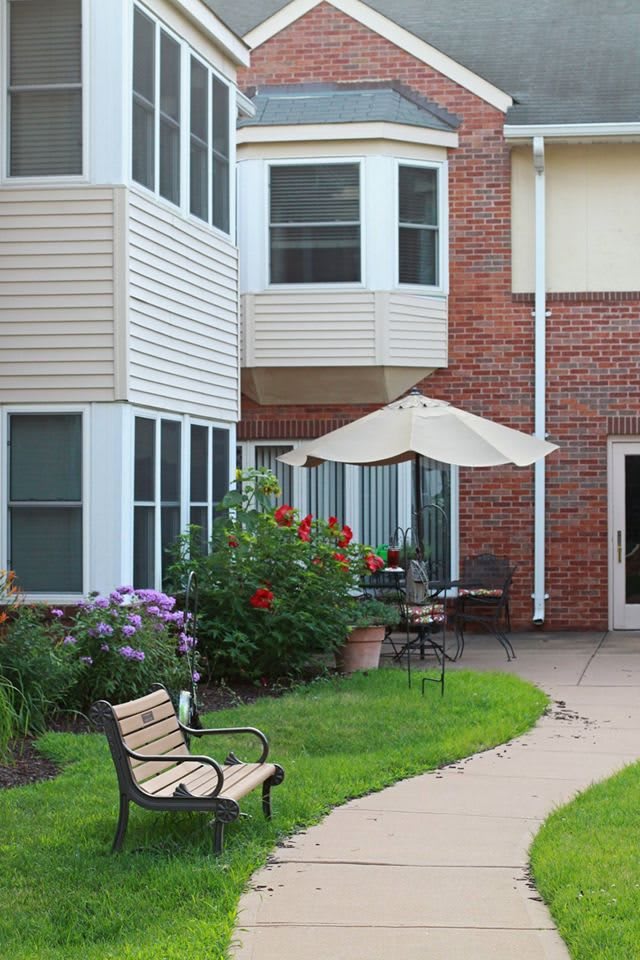 A cozy patio area with a garden, bench, and umbrella next to a brick building.