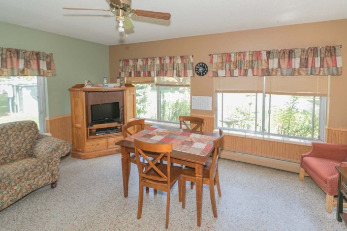 A cozy dining room with a wooden table and chairs, carpeted floor, and large windows adorned with curtains, featuring a ceiling fan and a fireplace in the corner.