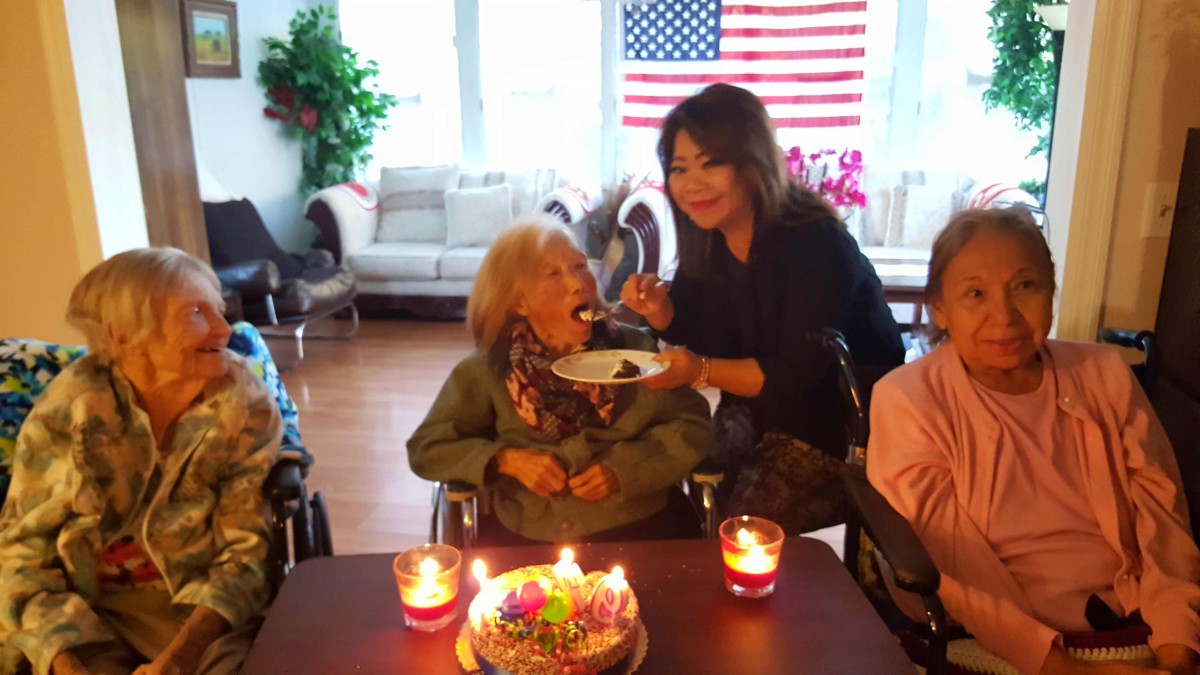An elderly woman celebrating her birthday with cake and candles, surrounded by friends, under an American flag.