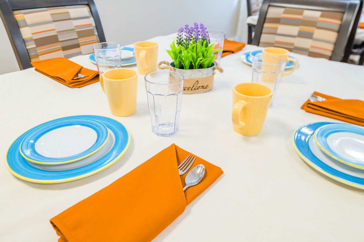 A neatly arranged dining table with orange napkins, blue and white plates, yellow cups, and a decorative centerpiece.