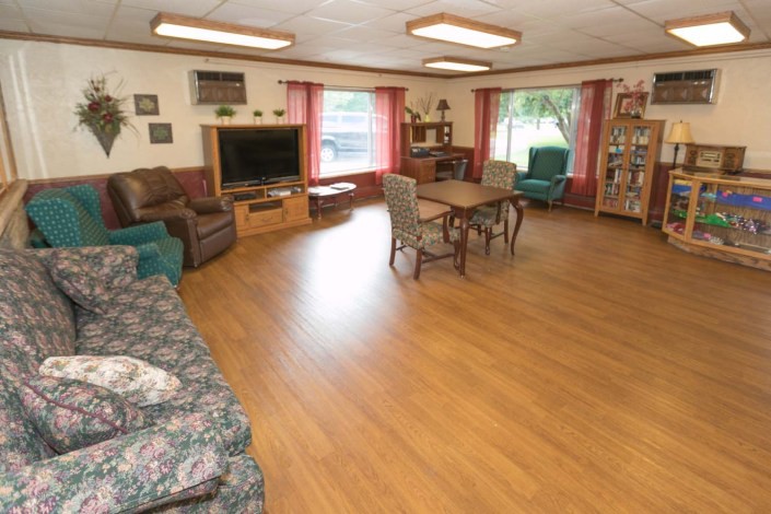 A cozy living room with wooden flooring, featuring a television, sofa, chairs, and a bookshelf.
