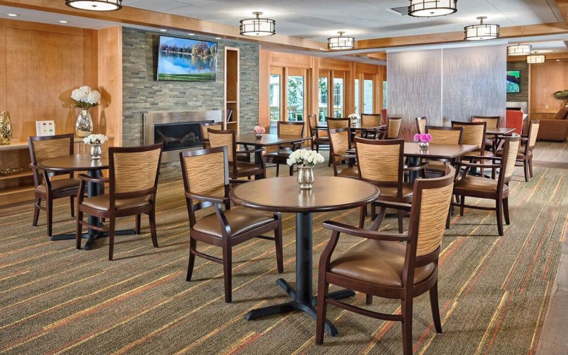 A cozy dining area with wooden tables and chairs arranged neatly, featuring a fireplace and a TV on the wall.