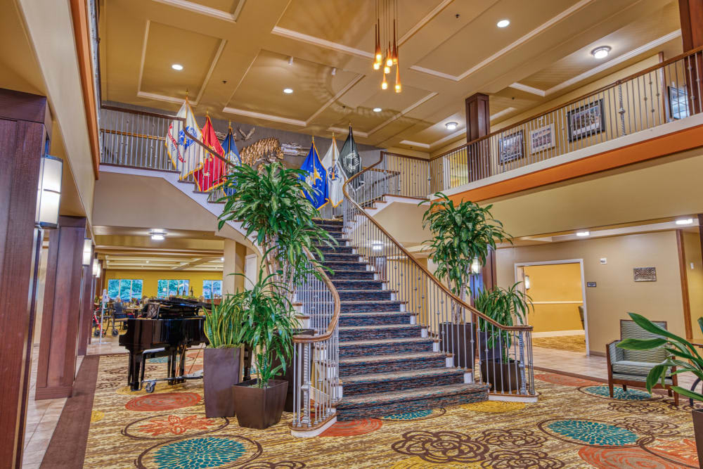 A spacious hotel lobby featuring a grand staircase, adorned with various flags and lush plants.