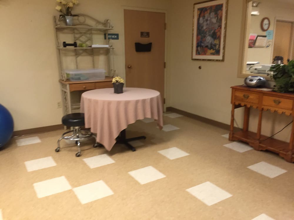 A quiet waiting room with a round table covered by a pink tablecloth, a chair, decorative plants, and artwork on the walls.