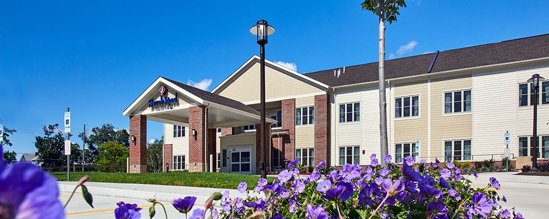 A modern two-story building with a covered entrance, surrounded by purple flowers under a clear blue sky.