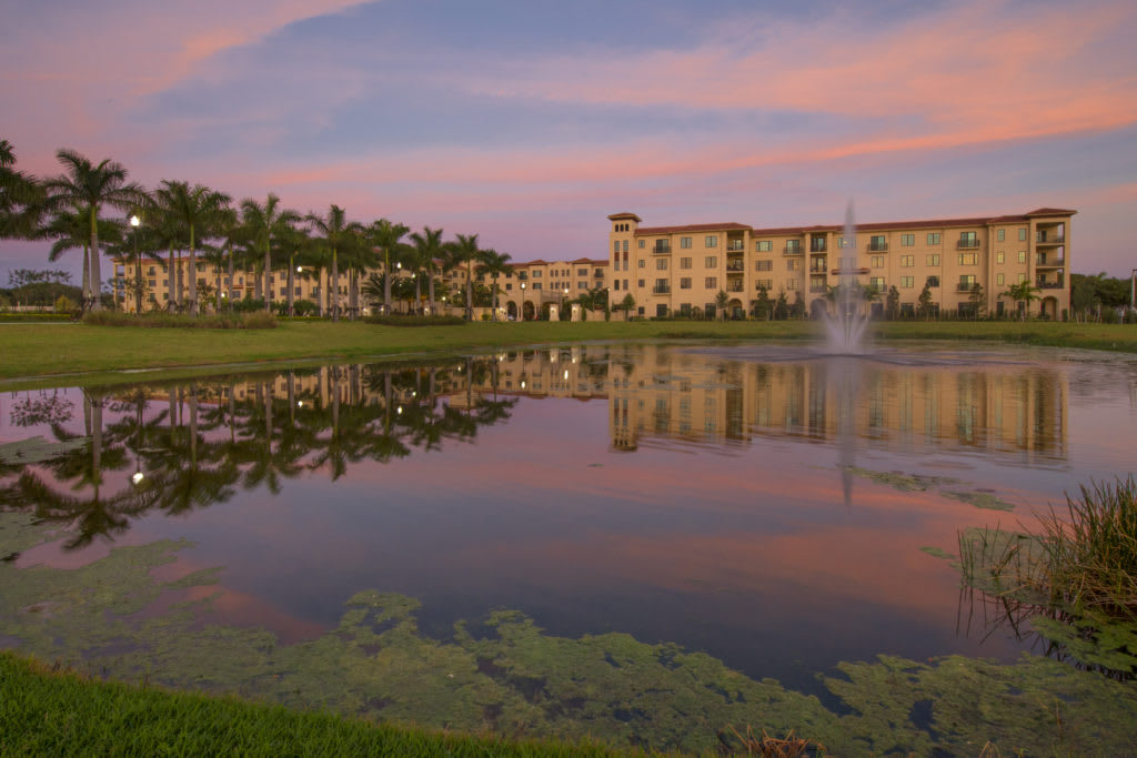 A serene landscape with a building and palm trees reflecting in a lake at sunset.