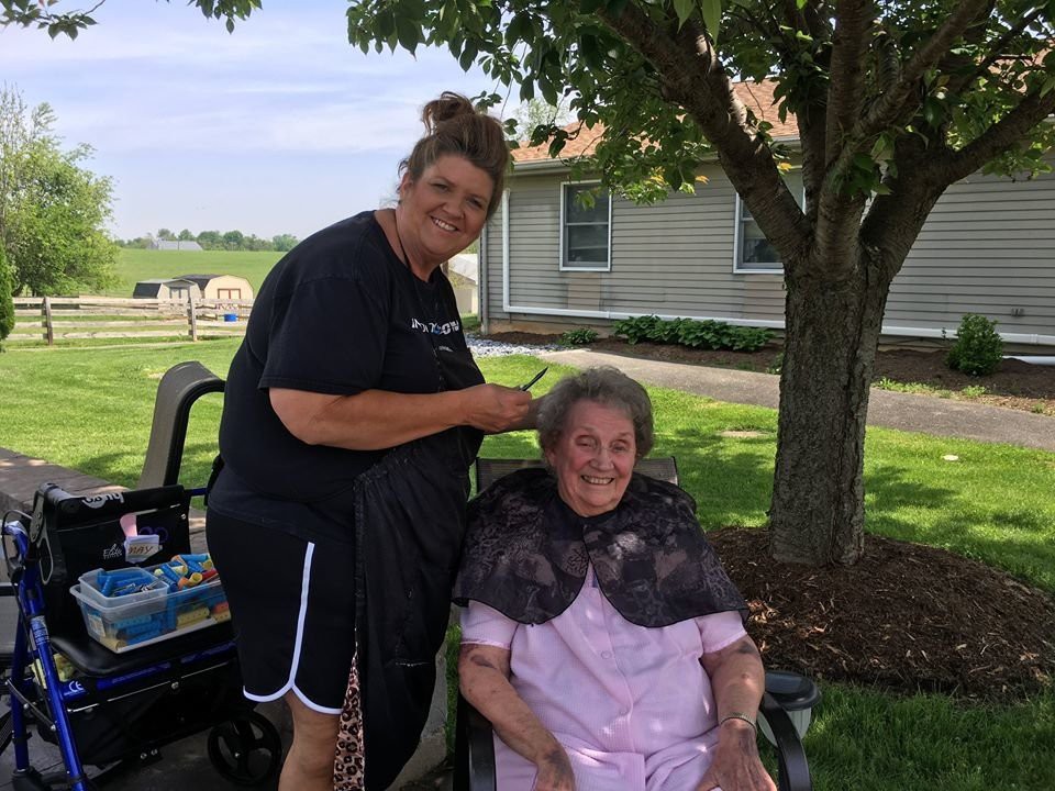 A woman giving a haircut to an elderly person under a tree outdoors.