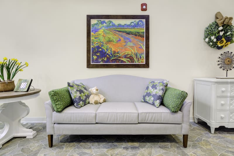 A cozy living room setup featuring a light gray sofa with decorative cushions, accompanied by a vibrant landscape painting on the wall.