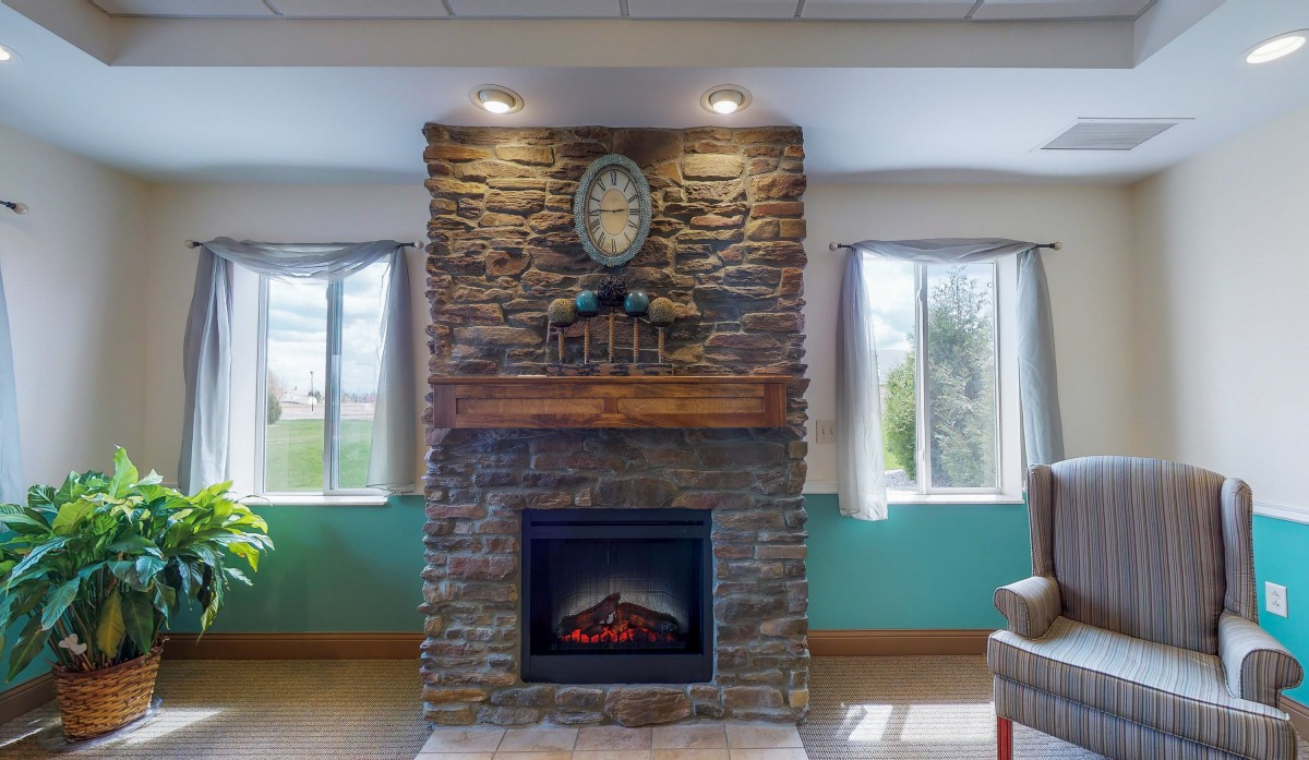 A cozy stone fireplace with a wooden mantle, flanked by two windows in a well-lit room.