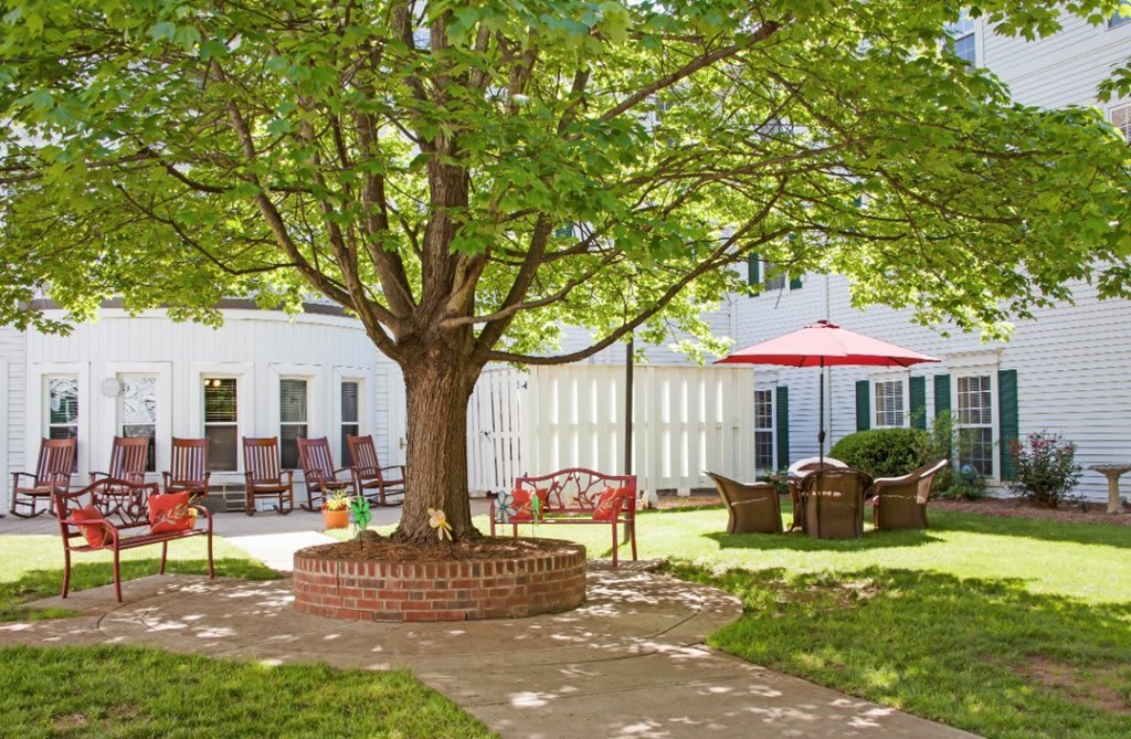 A serene backyard patio area featuring a large tree with a circular brick seating area, surrounded by outdoor furniture and a red sun umbrella.