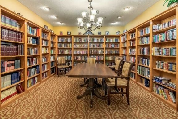A cozy library room with bookshelves lining the walls and a large wooden table surrounded by chairs under a chandelier.