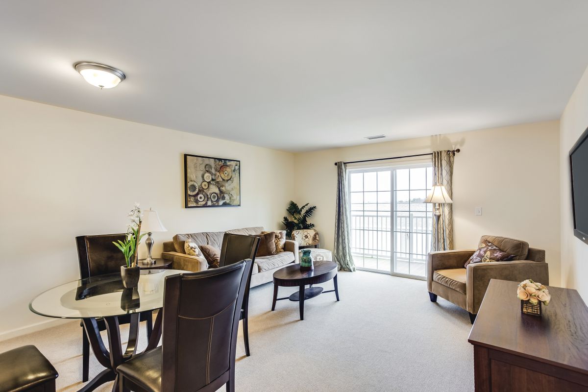 A well-furnished living room with a sofa, chairs, and a dining table, complemented by artwork on the wall and large windows allowing natural light.