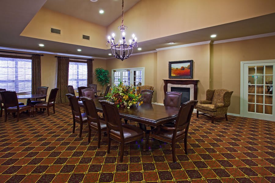 A well-furnished dining or conference room featuring a large table with chairs, a chandelier, a fireplace, and decorative elements such as a painting and flowers.
