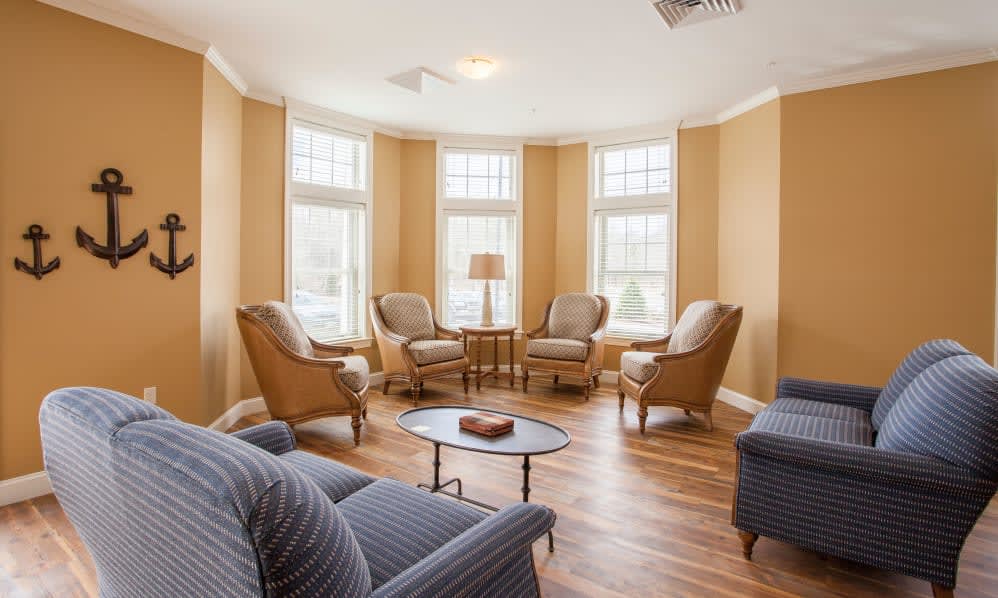 A cozy living room with armchairs arranged around a central table, illuminated by natural light from large windows.
