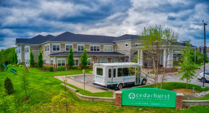 A modern building with a bus parked outside, surrounded by green landscaping and a cloudy sky.