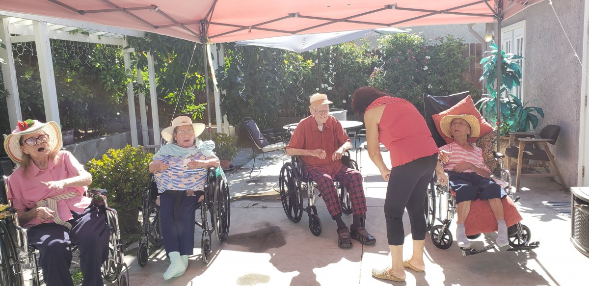 Two seniors in wheelchairs enjoy a sunny day under a canopy with their caregivers.