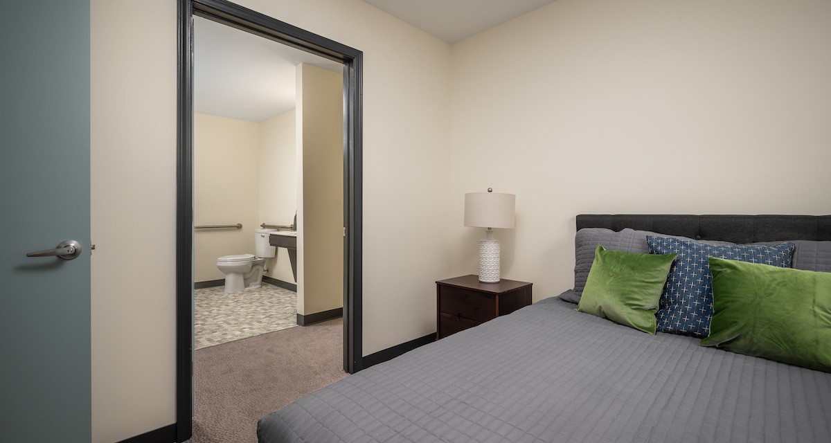 A cozy bedroom with a gray bedspread, a nightstand with a lamp, and an adjoining bathroom.